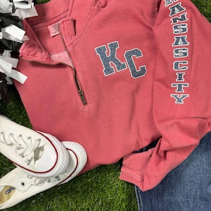 May include: A red sweatshirt with a white zipper and the letters "KC" in blue glitter on the front. The sleeve has the word "KANSAS CITY" in white glitter.