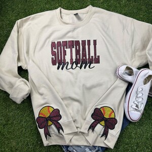 May include: A beige sweatshirt with the text "Softball Mom" in maroon glitter. The sleeves have maroon bows with a yellow softball on each.