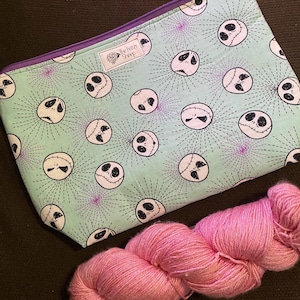 K&ouml;nnte beinhalten: Eine gr&uuml;ne Rei&szlig;verschlusstasche mit einem sich wiederholenden Muster von Jack Skellingtons Gesicht aus Nightmare Before Christmas. Die Tasche liegt auf einer braunen Oberfl&auml;che neben einer Spule pinkfarbenen Garns. Die Tasche hat einen violetten Rei&szlig;verschluss und ein kleines wei&szlig;es Etikett mit der Aufschrift "The Fuzzy Sheep".