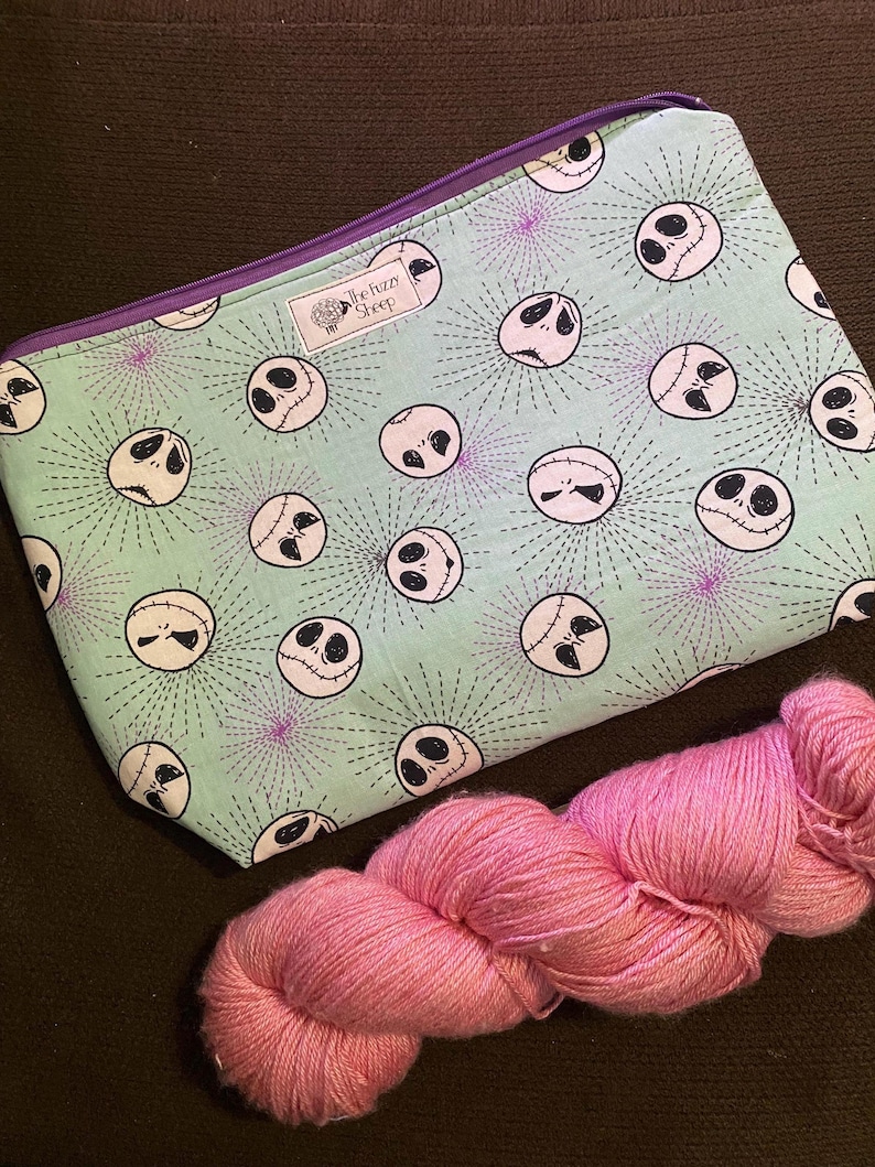 K&ouml;nnte beinhalten: Eine gr&uuml;ne Rei&szlig;verschlusstasche mit einem sich wiederholenden Muster von Jack Skellingtons Gesicht aus Nightmare Before Christmas. Die Tasche liegt auf einer braunen Oberfl&auml;che neben einer Spule pinkfarbenen Garns. Die Tasche hat einen violetten Rei&szlig;verschluss und ein kleines wei&szlig;es Etikett mit der Aufschrift "The Fuzzy Sheep".