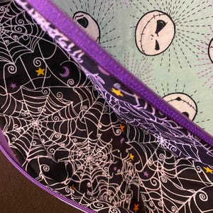 K&ouml;nnte beinhalten: Eine violette Rei&szlig;verschlusstasche mit einem schwarzen und wei&szlig;en Spinnennetz-Muster auf der Innenseite und einem gr&uuml;nen Stoff mit einem sich wiederholenden Muster von Jack Skellingtons Gesicht auf der Au&szlig;enseite.