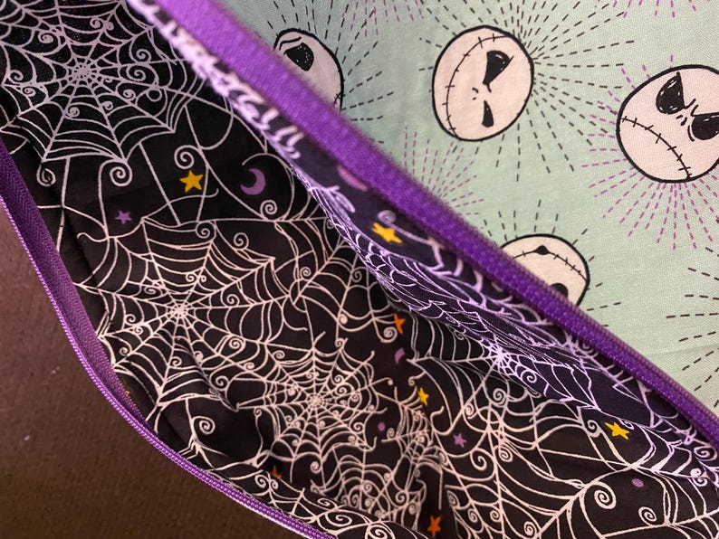K&ouml;nnte beinhalten: Eine violette Rei&szlig;verschlusstasche mit einem schwarzen und wei&szlig;en Spinnennetz-Muster auf der Innenseite und einem gr&uuml;nen Stoff mit einem sich wiederholenden Muster von Jack Skellingtons Gesicht auf der Au&szlig;enseite.