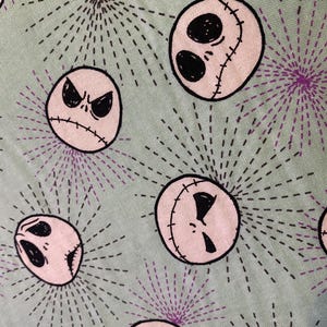 K&ouml;nnte beinhalten: Ein gr&uuml;ner Stoff mit einem sich wiederholenden Muster aus rosa und schwarzen Jack Skellington Gesichtern. Die Gesichter sind von einem schwarzen und violetten Sternenausbruchmuster umgeben.
