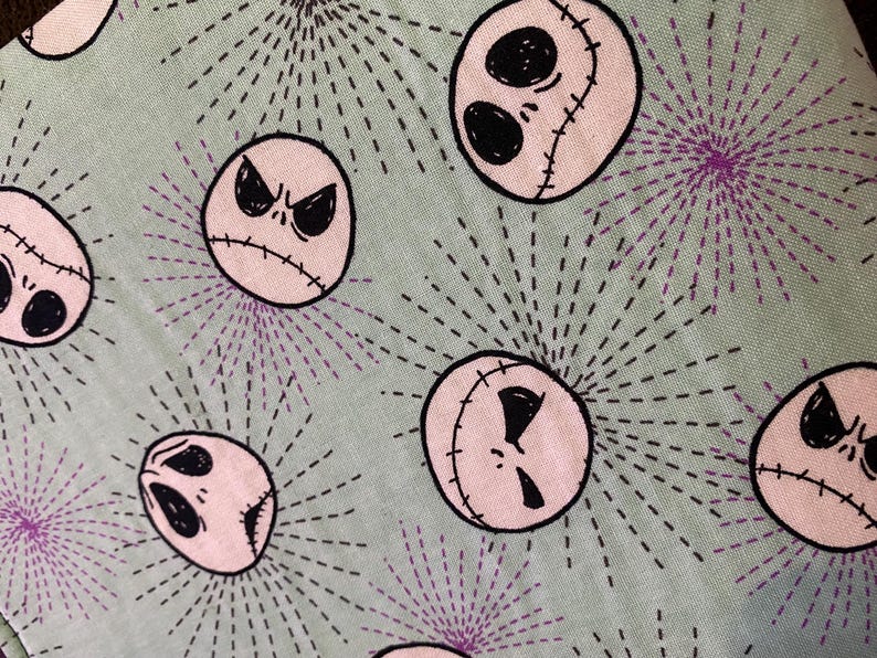 K&ouml;nnte beinhalten: Ein gr&uuml;ner Stoff mit einem sich wiederholenden Muster aus rosa und schwarzen Jack Skellington Gesichtern. Die Gesichter sind von einem schwarzen und violetten Sternenausbruchmuster umgeben.