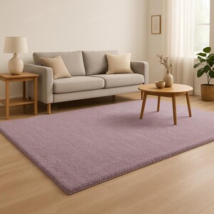 May include: A rectangular, lavender-colored rug with a textured surface. The rug is placed on a hardwood floor in a living room setting. A light gray sofa with beige pillows and a wooden coffee table are also visible.