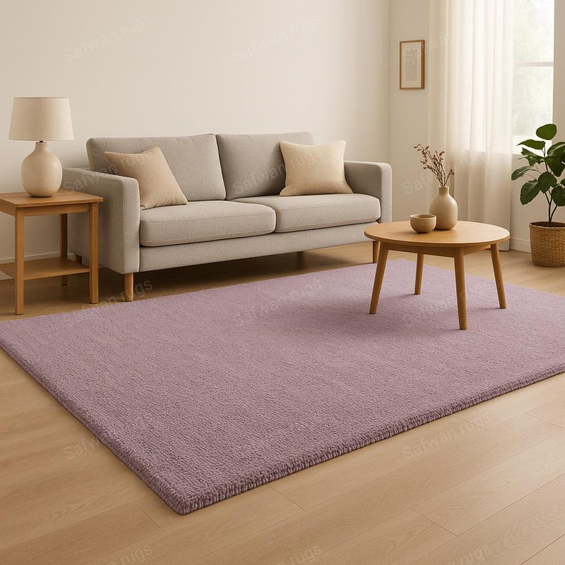May include: A rectangular, lavender-colored rug with a textured surface. The rug is placed on a hardwood floor in a living room setting. A light gray sofa with beige pillows and a wooden coffee table are also visible.