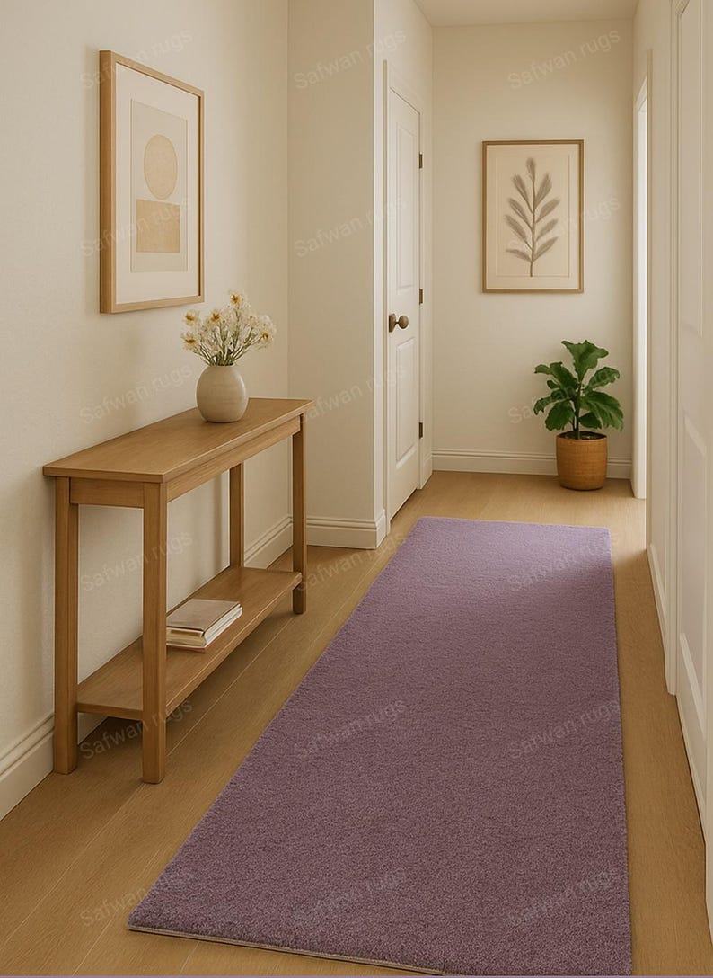 May include: A hallway with a light purple rectangular rug, a wooden console table, and framed artwork. The walls are painted a neutral color, and the floor is hardwood. A vase of flowers sits on the table, and a potted plant is visible.