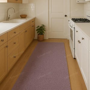 May include: A rectangular, mauve-colored rug in a kitchen setting. The rug is placed on a hardwood floor, extending from the base of the cabinets towards the center of the room. The kitchen features light-colored cabinets and a white door.