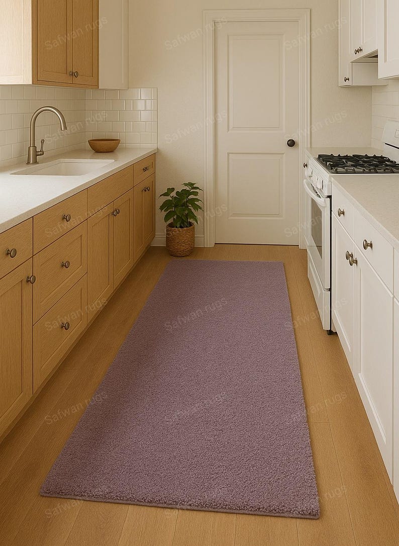 May include: A rectangular, mauve-colored rug in a kitchen setting. The rug is placed on a hardwood floor, extending from the base of the cabinets towards the center of the room. The kitchen features light-colored cabinets and a white door.