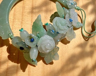 May include: A pale green jade bangle bracelet adorned with carved jade floral and leaf motifs. The flowers and leaves are attached to the bangle with light green cord and feature small blue thread accents at the center of each blossom.