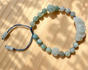 May include: A bracelet featuring light green jade beads, two carved floral beads, and a large curved carved floral centerpiece. The beads are strung on a blue cord with an adjustable sliding knot closure and two small cylindrical jade end beads.