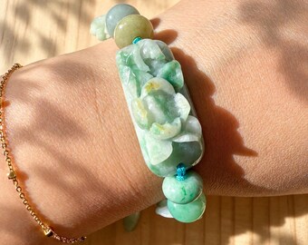 May include: A bracelet featuring a central rectangular green jade bead carved with a floral design, flanked by smaller round green jade beads on a blue cord, accompanied by a separate thin gold chain bracelet with small round gold beads.