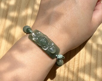 May include: A bracelet featuring a central rectangular dark green stone plaque carved with a floral motif. The plaque is flanked by smaller spherical dark green stone beads, all strung on a light green cord with adjustable knotted ends.