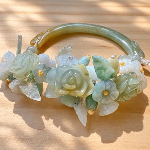 May include: A jade bangle bracelet adorned with carved light green and white stone flowers and leaves. The floral arrangement features small yellow centers and is attached to a smooth, curved jade band with a subtle yellow-green gradient.