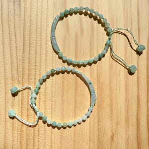 May include: Two jade bead bracelets with adjustable drawstrings. Each bracelet features small, round, light green beads and a curved jade accent piece. The bracelets are displayed on a light brown wooden surface.