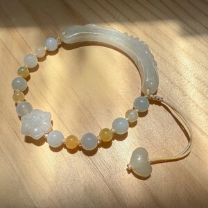 Könnte beinhalten: Hellgrünes Jade-Armband mit einem floralen Anhänger und einem gebogenen, geschnitzten Jade-Element. Das Armband hat runde, hellgrüne und gelbe Perlen sowie einen herzförmigen Jade-Anhänger am Ende.