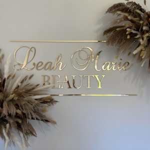 以下が含まれることがあります： 「Leah Marie BEAUTY」の文字がエレガントな筆記体とブロック体で書かれた、金色の鏡面サイン。サインは2本の水平な金色の線で縁取られ、明るい灰色の壁にドライフラワーアレンジメントで飾られています。