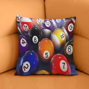 May include: A decorative pillow featuring a close-up of billiard balls. The balls are various colors, including red, orange, purple, and yellow, with white numbers. The pillow is on an orange surface.