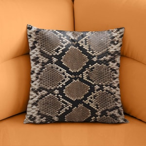 Peut inclure: Coussin carré décoratif avec un motif peau de serpent. Le coussin présente un motif répété d'écailles marron, noires et beiges. Le coussin est posé sur un canapé orange.