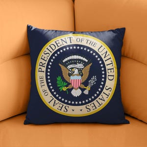 Pode incluir: Uma almofada quadrada azul marinho com o Selo Oficial do Presidente dos Estados Unidos. O selo é circular, com uma borda dourada e texto branco. O centro apresenta uma águia com um escudo, ramo de oliveira e flechas.