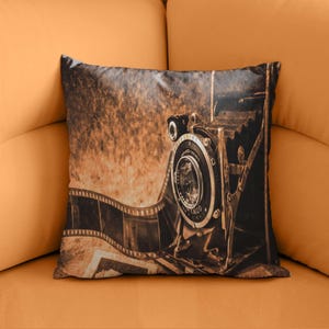 Puede incluir: Cojín decorativo con una impresión en tonos sepia de una cámara vintage y una tira de película. El diseño evoca una sensación retro, con los detalles de la cámara y las curvas de la tira de película. El fondo tiene un aspecto envejecido y texturizado.
