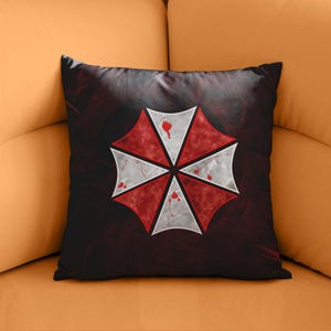 May include: A black and red throw pillow featuring a white umbrella logo with blood splatter. The umbrella has six points and is set against a dark red background.