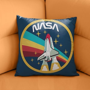 Puede incluir: Un cojín cuadrado azul marino con el logotipo de la NASA y un diseño de transbordador espacial. El diseño incluye un círculo amarillo, estrellas y rayas de colores. El cojín está colocado en un sofá naranja.
