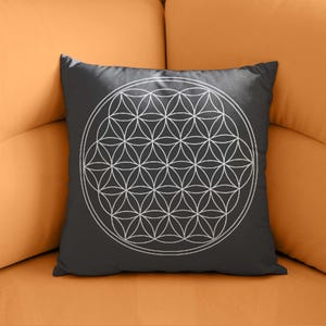 May include: A square, dark gray throw pillow featuring a white, geometric Flower of Life pattern. The design is a series of overlapping circles forming a symmetrical floral shape. The pillow is resting on an orange couch.