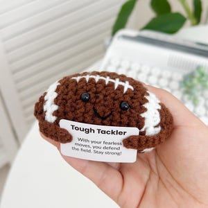 Op de afbeelding: Een gehaakt pluche voetbal speelgoed dat een kaart vasthoudt met de tekst "Tough Tackler With your fearless moves, you defend the field. Stay strong!"