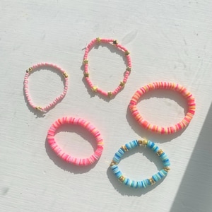 May include: Five colorful beaded bracelets. The bracelets feature pink, orange, yellow, and blue beads. Some bracelets have gold star or round accents. The bracelets are arranged on a white surface.