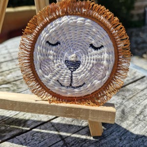 Peut inclure: Un dessous de verre rond tissé représentant un visage de lion. Le lion a les yeux fermés, un nez noir et une bouche cousue. Le dessous de verre a une frange marron clair et est posé sur un petit chevalet en bois.