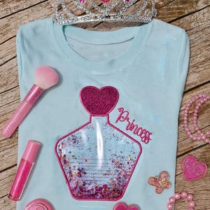 Peut inclure: T-shirt bleu clair avec un motif de flacon de parfum scintillant et le mot "Princess". Les accessoires comprennent un diadème, un pinceau de maquillage, un brillant à lèvres et des ornements en forme de cœur. Thème princesse.