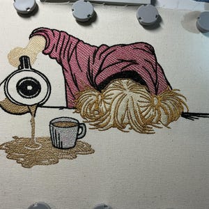 Puede incluir: Diseño bordado que representa a una persona con la cabeza gacha, derramando café de una jarra en una taza y sobre la superficie. La persona lleva una camisa rosa. El pelo es rubio con dos coletas pequeñas. El café y la jarra son marrones y blancos.