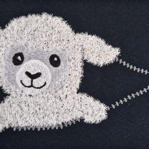 Puede incluir: Cordero blanco bordado con ojos negros y una cremallera unida a su espalda. El cordero está sobre un fondo azul oscuro.