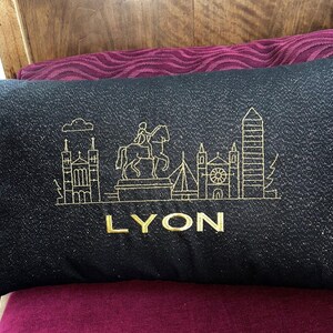 May include: Black rectangular pillow with gold embroidered skyline of Lyon, France. The pillow features a detailed outline of buildings, a statue, and the word "LYON" in gold lettering. The pillow is resting on a burgundy surface.
