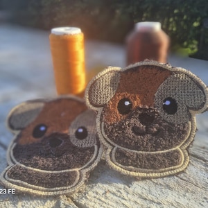 Puede incluir: Dos parches bordados con forma de perro, con detalles en marrón, beige y negro. Los parches tienen un aspecto texturizado y están sobre un fondo borroso con carretes de hilo naranja. Un artículo artesanal lindo.
