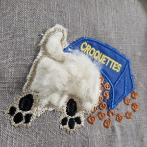 Puede incluir: Parche bordado que representa la parte trasera de un perro con almohadillas negras y una cola blanca y esponjosa. Una bolsa azul con la inscripción "CROQUETTES" derrama croquetas marrones y circulares. Sobre un fondo de tela gris.