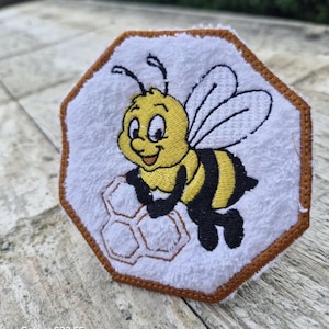 Peut inclure: Écusson blanc brodé avec un dessin d'abeille de dessin animé. L'abeille est jaune et noire, souriante et tient un nid d'abeilles. L'écusson a une bordure marron et une forme octogonale.