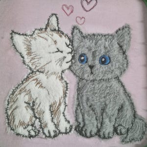 以下が含まれることがあります： ピンク色の背景に、2匹のふわふわした子猫が刺繍されています。白い子猫が青い目の灰色の猫にキスをしています。3つのピンクのハートが上に浮かんでいます。子猫は、質感のあるぬいぐるみのような効果でステッチされています。