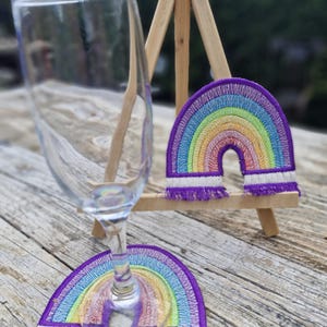 Könnte beinhalten: Ein klares Weinglas steht auf einer Holzoberfläche neben zwei regenbogenförmigen, bestickten Untersetzern. Die Untersetzer haben ein farbenfrohes Regenbogenmuster mit lila Fransen und einem weißen Rand. Eine kleine Holzstaffelei hält einen Untersetzer.