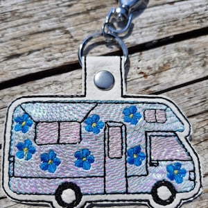 Peut inclure: Un porte-clés en forme de camping-car avec un dos blanc et un fermoir argenté. Le camping-car est violet clair avec des broderies de fleurs bleues. Les roues et les fenêtres sont noires. Le porte-clés est sur une surface en bois.