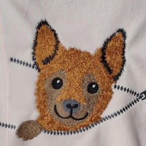 Può includere: Tessuto rosa chiaro con un disegno di cane ricamato. Il cane è marrone con dettagli neri e sembra emergere da una cerniera grigia e nera.