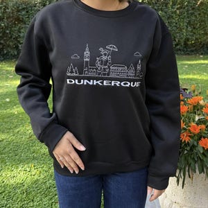 Peut inclure: Sweat-shirt noir avec un dessin blanc de la skyline de Dunkerque. Le motif comprend des bâtiments, une personne avec un parapluie et le mot "DUNKERQUE". Le sweat-shirt a des manches longues.