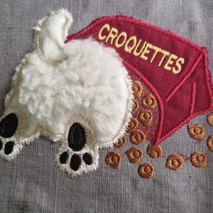 Puede incluir: Parche bordado con la parte trasera de un perro blanco y esponjoso con almohadillas negras y una bolsa roja de "CROQUETTES" con croquetas marrones derramadas. El parche está sobre un fondo de tela gris.