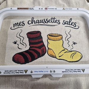 Peut inclure: Un cercle à broder maintient une toile avec l'expression « mes chaussettes sales » et un dessin animé de deux chaussettes malodorantes. L'une est rayée rouge et noire, l'autre est jaune. Des mouches et des lignes ondulées suggèrent l'odeur des chaussettes.