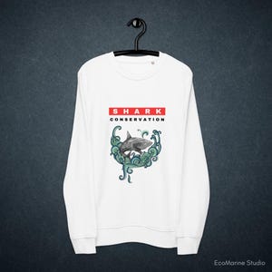 Könnte beinhalten: Weißes Sweatshirt mit einem geometrischen Hai-Design, umgeben von türkisfarbenen Wellen. Der Text "SHARK CONSERVATION" ist in Rot und Schwarz über dem Hai-Motiv gedruckt. Das Sweatshirt hängt an einem schwarzen Kleiderbügel.