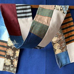 Peut inclure: Une longue écharpe rectangulaire en patchwork composée de divers panneaux de tissu. Le motif comprend des sections unies bordeaux, bleues et beiges, ainsi que des segments à rayures noires et blanches, des motifs floraux bruns et des lignes horizontales multicolores.