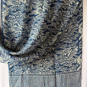 Peut inclure: Une écharpe rectangulaire présentant un fond bleu profond avec un motif botanique répétitif blanc cassé composé de feuilles et de baies. Le bord inférieur affiche une bordure distincte avec des rayures verticales et un petit détail en pointillés.