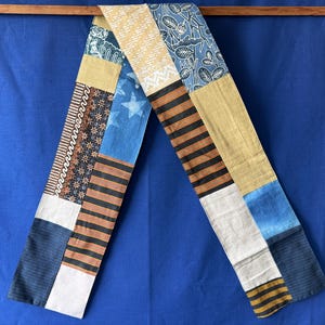 Peut inclure: Une longue écharpe rectangulaire en patchwork composée de divers motifs de tissus, notamment du bleu avec des étoiles blanches, des rayures horizontales noires et orange, du jaune moutarde uni, du blanc, du bleu foncé et un imprimé floral bleu.