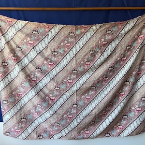 Op de afbeelding: Een rechthoekige wikkelrok met een traditioneel batikpatroon met diagonale banen van witte ingewikkelde motieven afgewisseld met bloemmotieven in tinten oudroze, bruin en groenblauw. De stof is voorzien van strikbanden aan de bovenste hoeken.